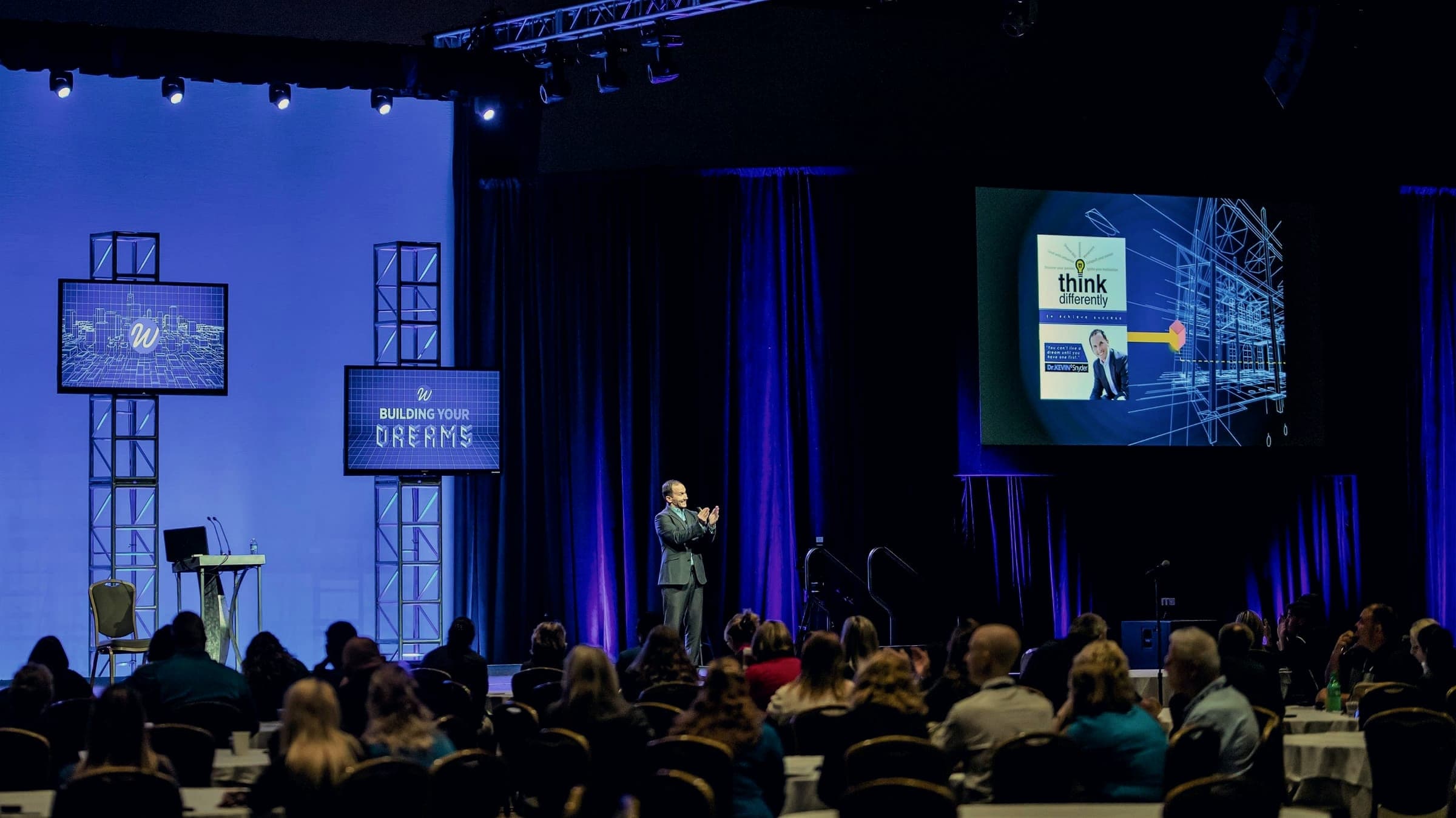 Kevin speaking on a large blue-lit conference stage with three screens behind him