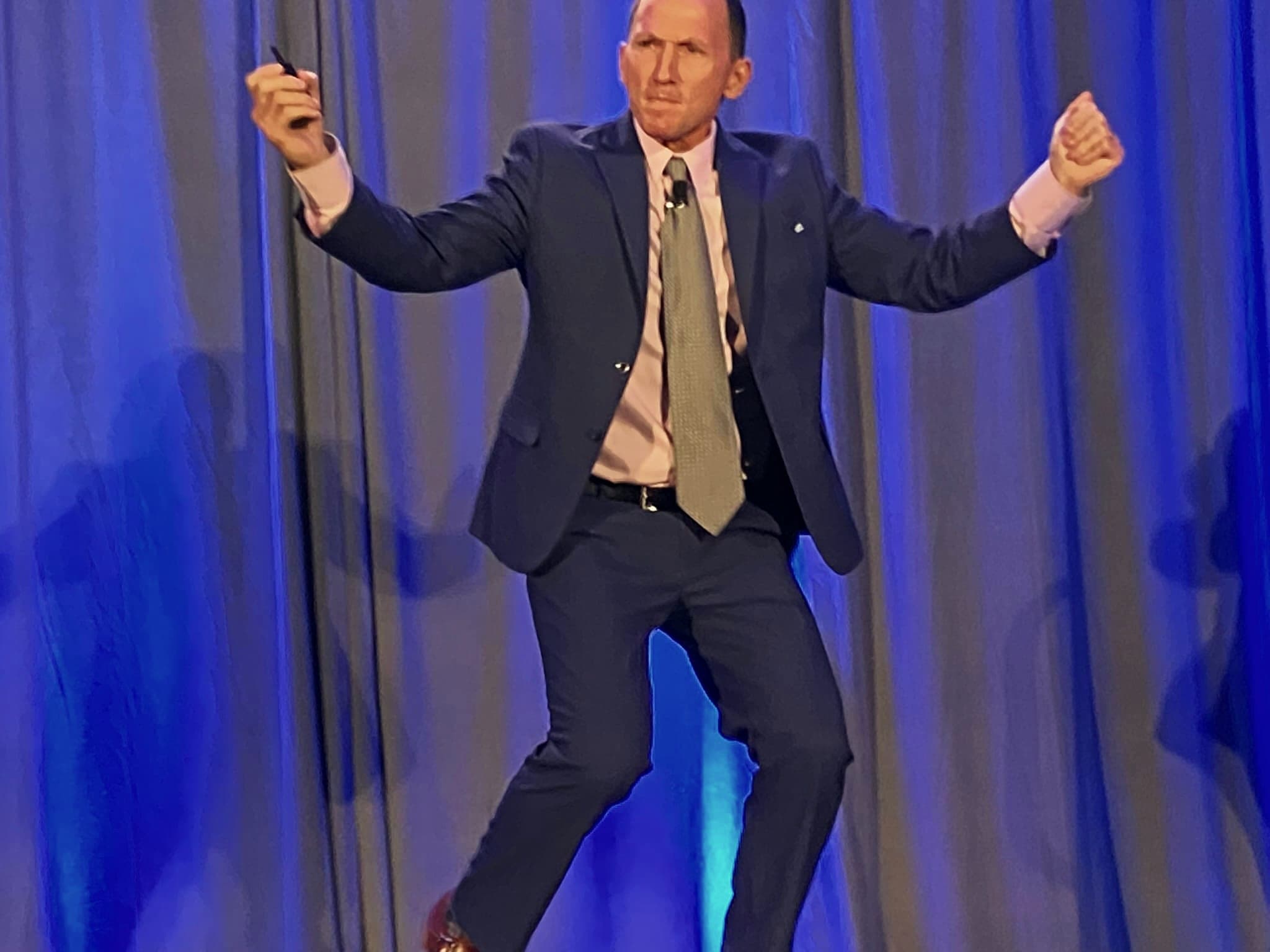 Kevin on stage in front of a blue curtain, arms out mid-story