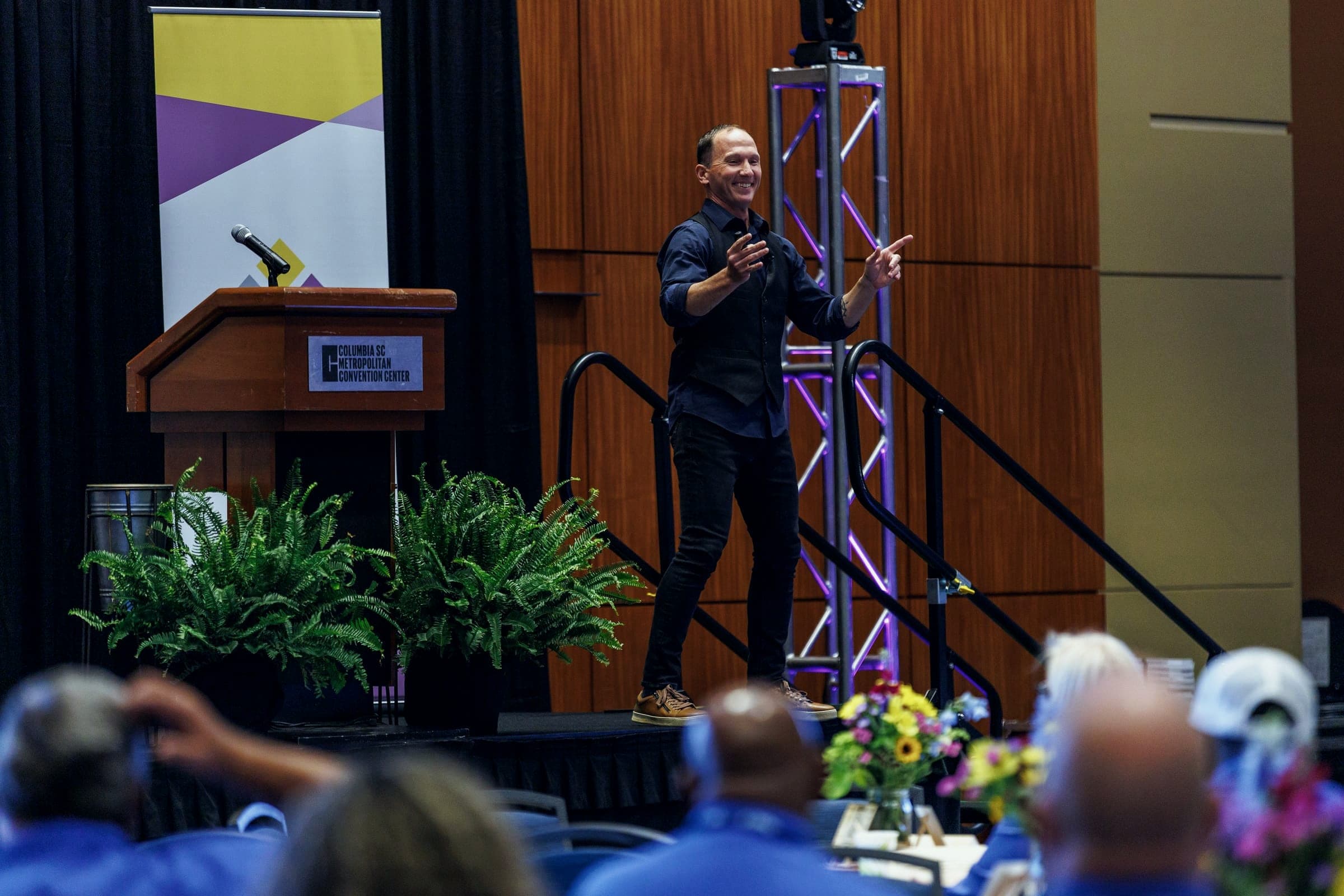 Kevin speaking on a stage framed by lighting truss, audience in the foreground