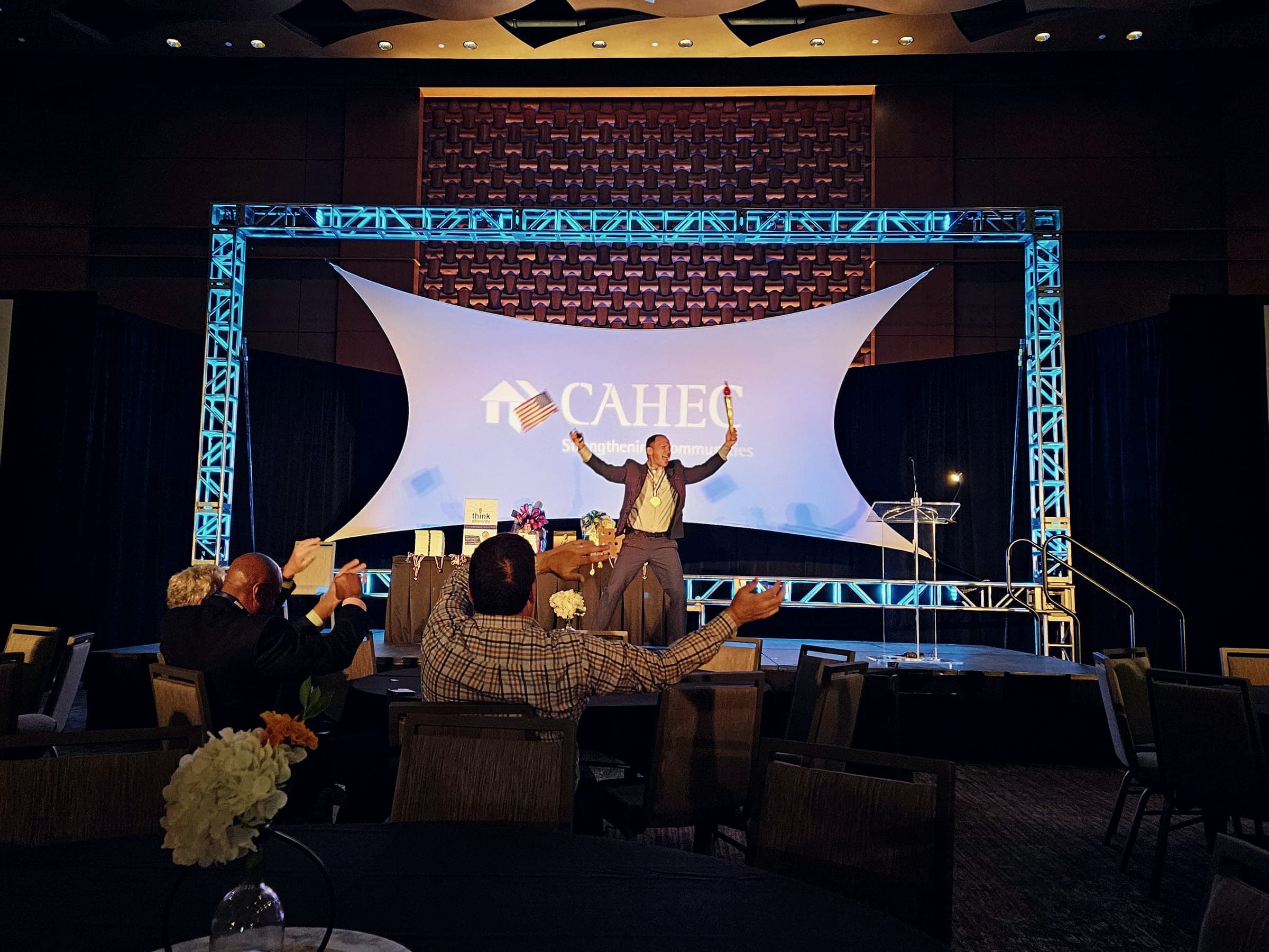 Kevin waving from a stage with a large lit backdrop as the audience waves back